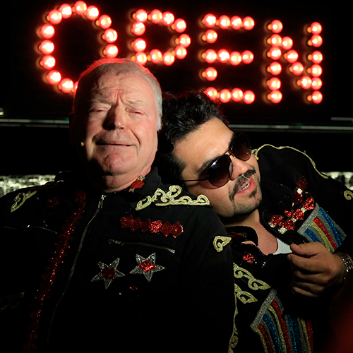 Luis leaning on a fellow actor under a glowing ‘OPEN’ sign in Mahagonny.