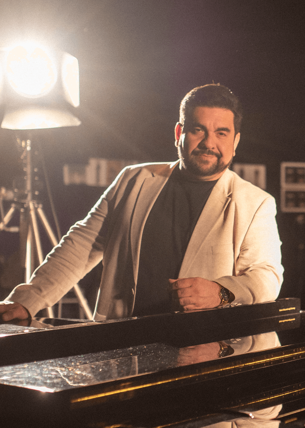 A spotlight shines behind Luis in a dark setting, and she stands with her arms crossed on a grand piano, wearing a black T-shirt and white jacket.